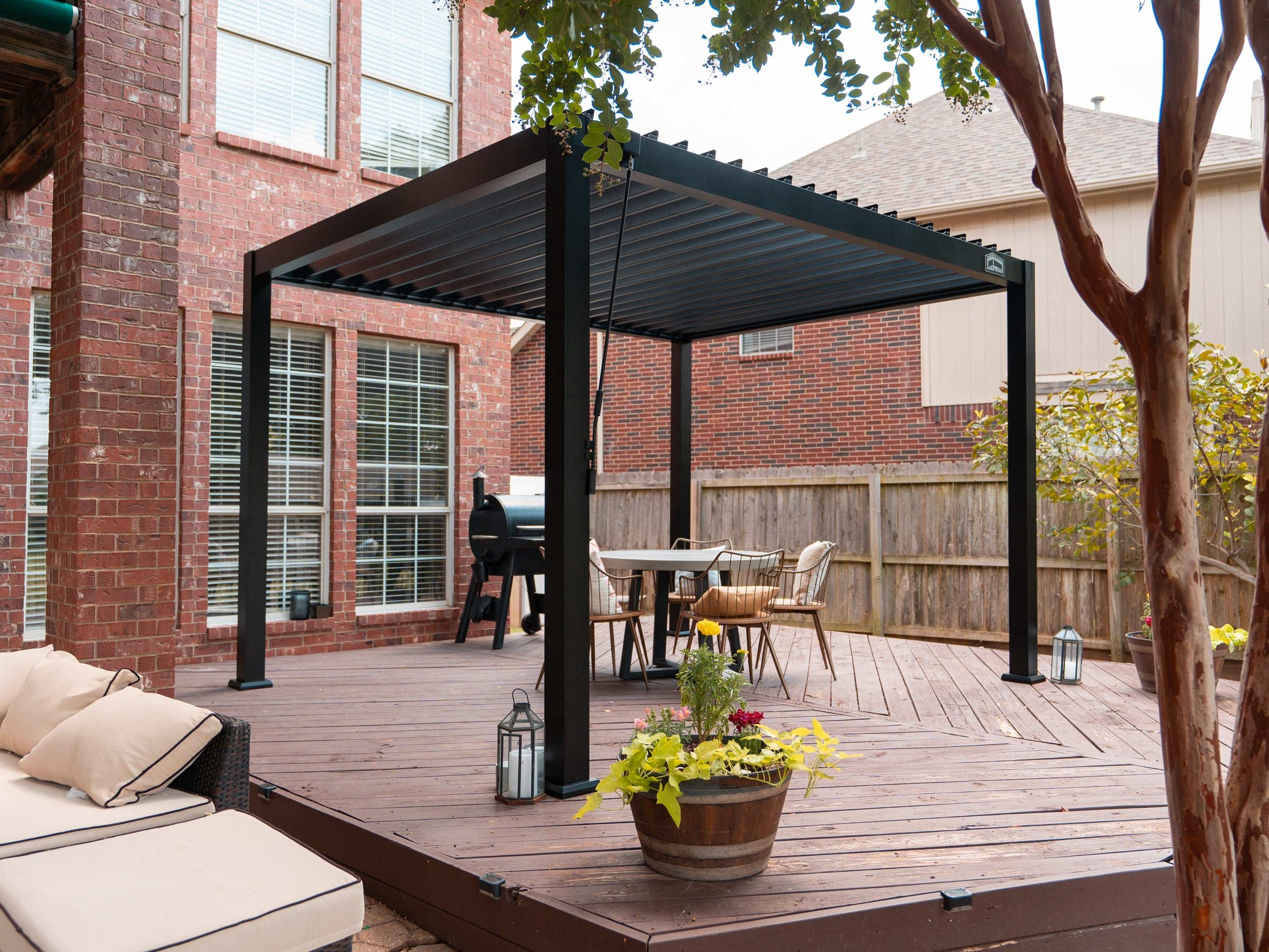A black louvered pergola over a dining set and grill on a wooden deck next to a brick house.