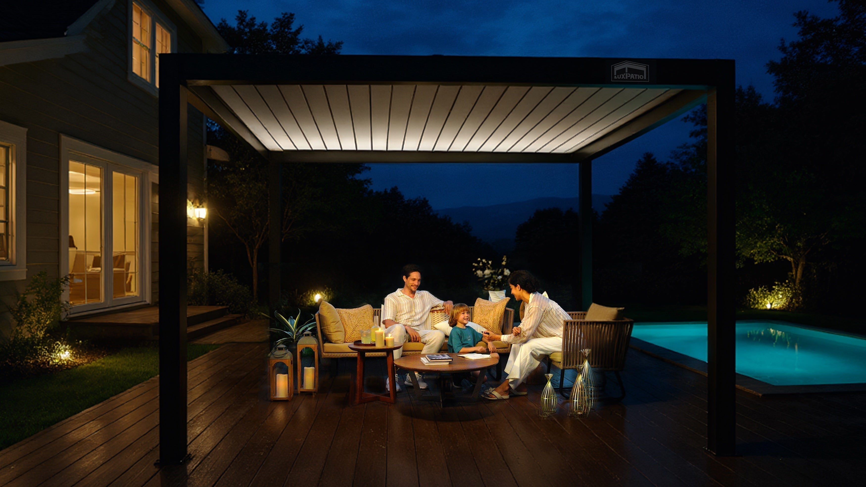 A family of three relaxes together on outdoor furniture under a lighted pergola at night. The scene is set on a  aluminum patio next to a swimming pool.