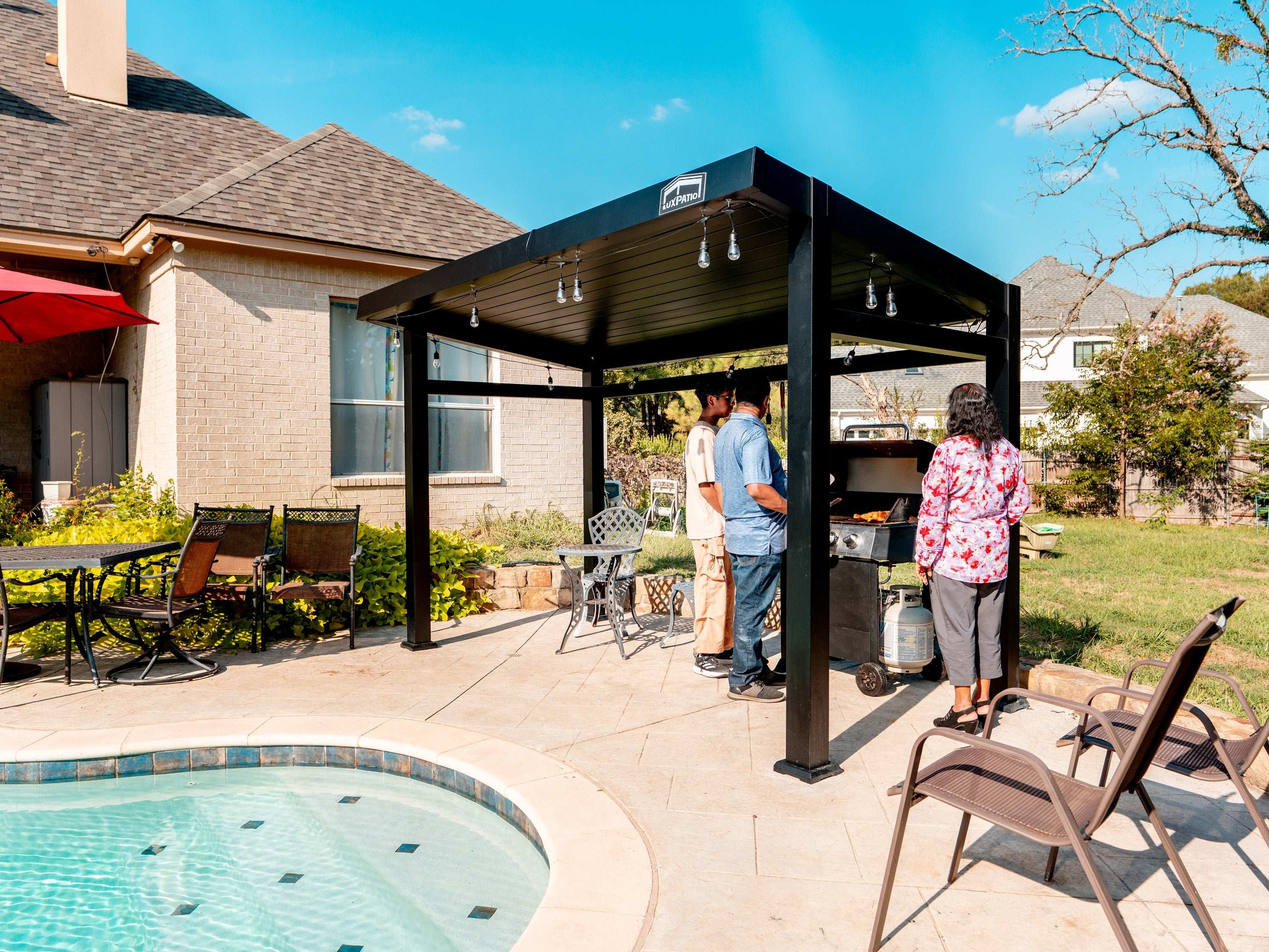 People grilling under a black metal gazebo with string lights next to a pool in a backyard.